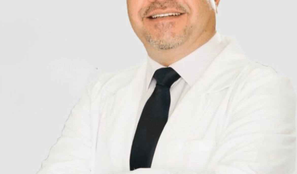 Smiling male dentist wearing glasses, a white coat, and a black tie, standing with arms crossed against a light background